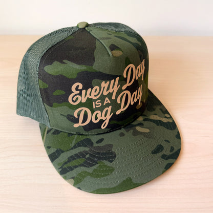 A dark green camouflage trucker cap with a curved brim and mesh back, featuring the text 'Every Day is a Dog Day' in tan script on the front panel, resting on a light wood surface.