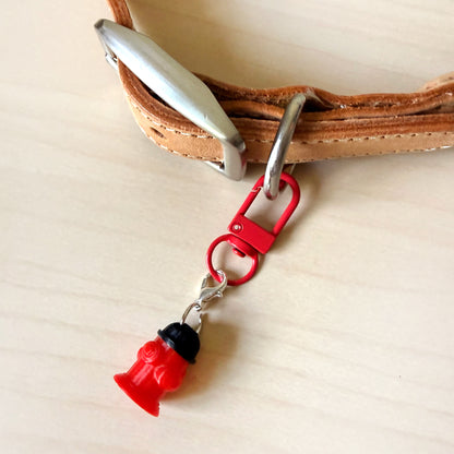 A close-up of a tan leather strap with a silver metal buckle and a red carabiner clip attached, which holds a small red and black fire hydrant charm, all resting on a light wood surface.