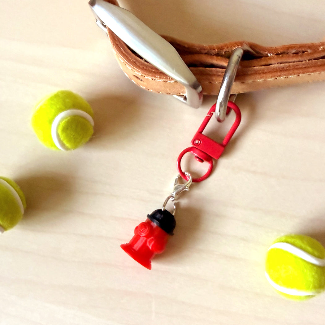 A red firefighter helmet-shaped keychain or bag charm with a silver clasp, shown attached to a brown leather strap on a light wood surface, with tennis balls as props.