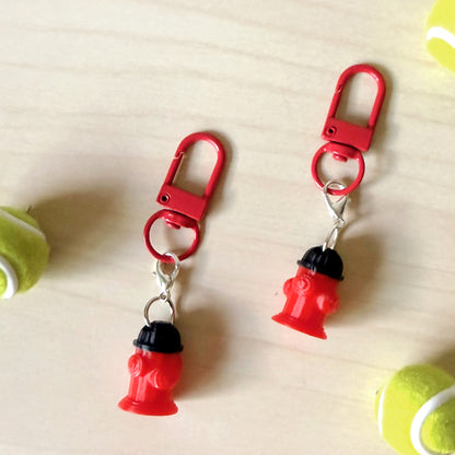 A pair of red fire hydrant keychains with red carabiners and silver jump rings, displayed on a light wood surface with tennis balls in the corners.