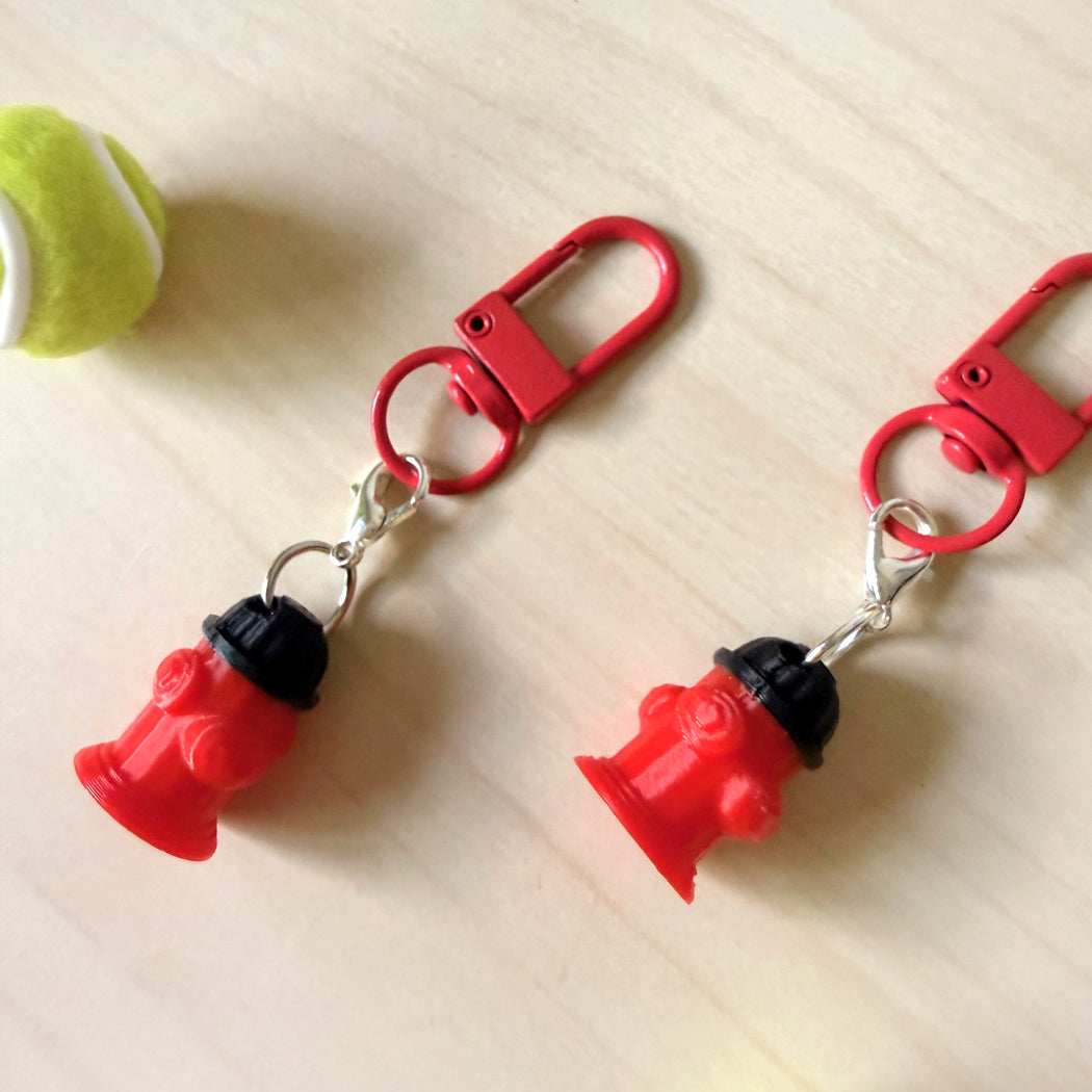 Two red fire hydrant keychains with silver carabiners, displayed on a light wood surface next to a yellow tennis ball.