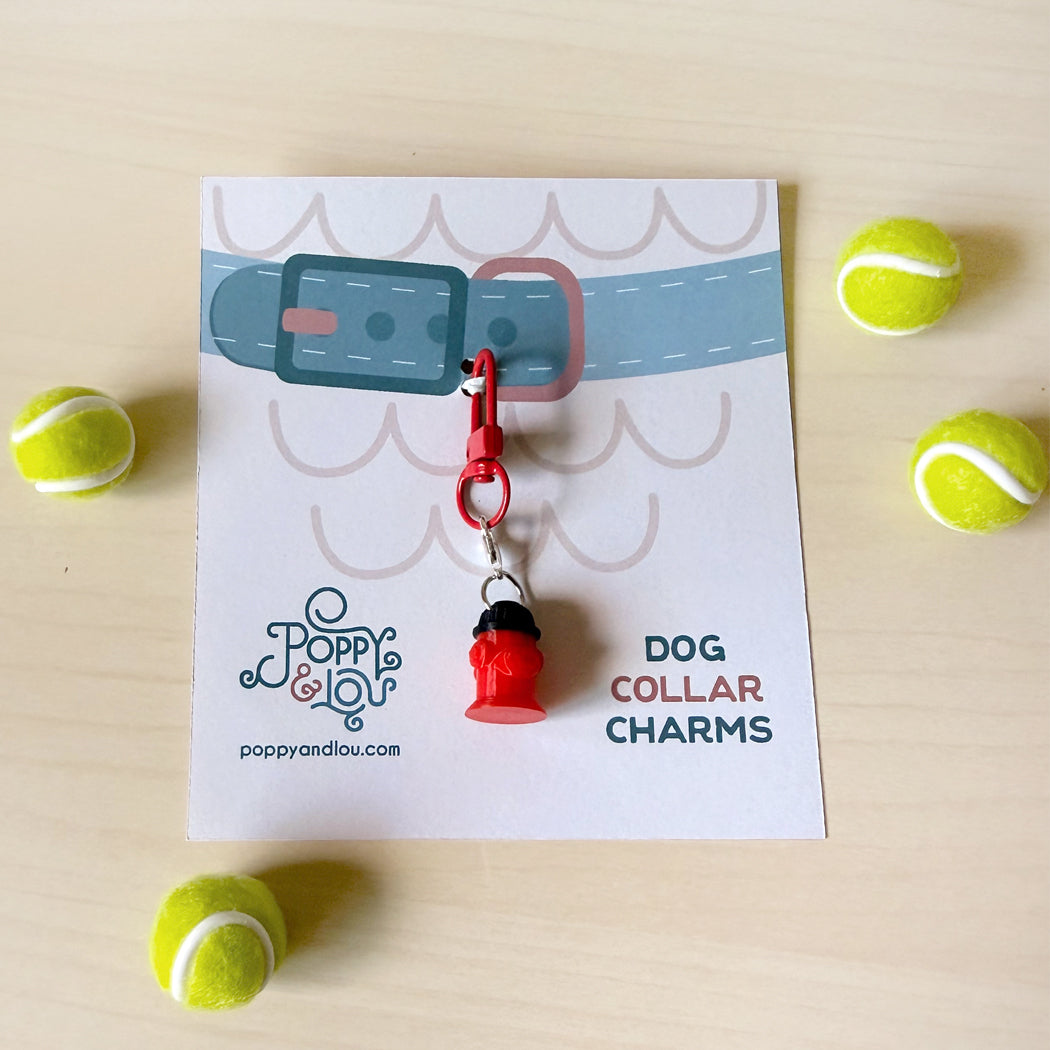 A red bell-shaped dog collar charm with a silver clasp, attached to a white printed card that reads 'DOG COLLAR CHARMS' and shows the 'Poppy and Lou' brand logo. The card is laid on a light wood surface and surrounded by four yellow tennis balls.