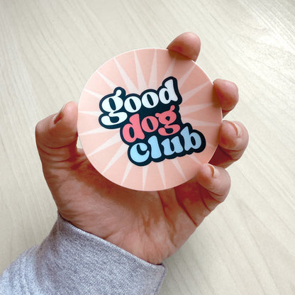 Hand holding a pink round Good Dog Club vinyl sticker with retro lettering and sunburst design.