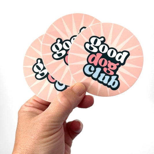 Hand holding a pink round Good Dog Club vinyl sticker with retro lettering and sunburst design.