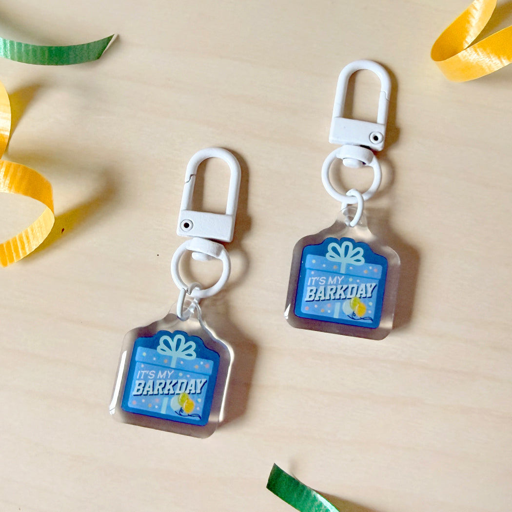 Two acrylic keychain charms shaped like gift boxes with white clasp loops, printed with 'IT'S MY BARKDAY' in blue and yellow, displayed on a light wood surface with colorful paper ribbons.