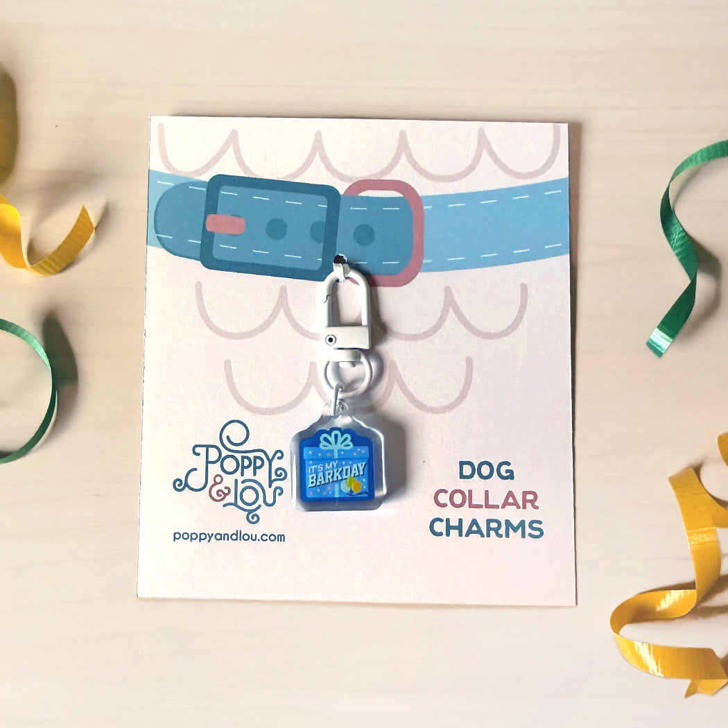A blue and white embroidered dog collar charm by Poppy & Lou, attached to a silver clasp and displayed on its branded backing card. The card features a graphic of a blue collar with a pink buckle, set against a light wood background with decorative green and yellow ribbons.