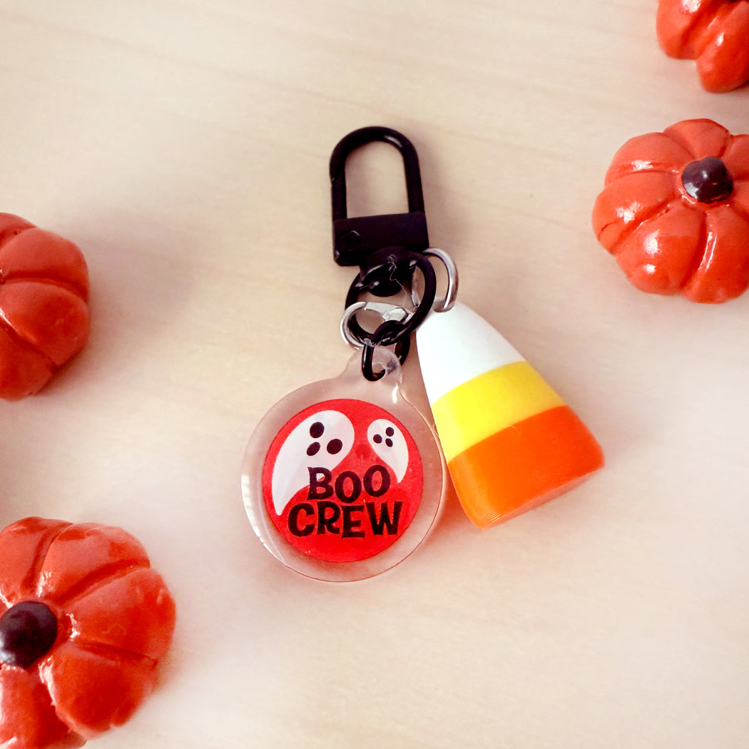 A Halloween-themed keychain featuring a clear round charm with a red and white ghost and the words 'BOO CREW' in black, paired with a candy corn charm, on a light wood background with small orange pumpkin decorations.