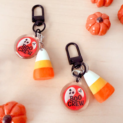 Two Halloween-themed keychains on a light wood background, styled with small orange pumpkins. The keychains feature a black lobster clasp, a candy corn charm, and a round clear charm with a ghost graphic and the words 'BOO CREW'.