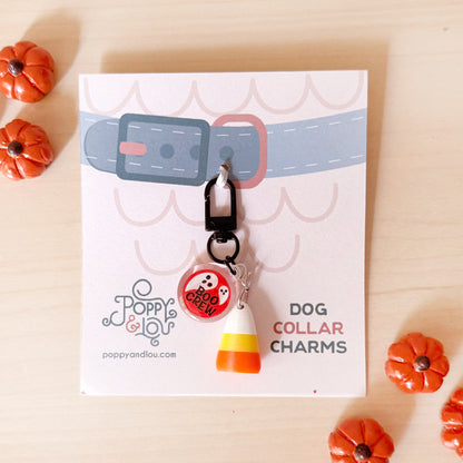 A dog collar charm set displayed on a branded card, featuring a black carabiner clip with a round red-and-white 'BOO HOO MEOW' charm and a candy corn charm, photographed on a light wood surface with small orange pumpkin props.