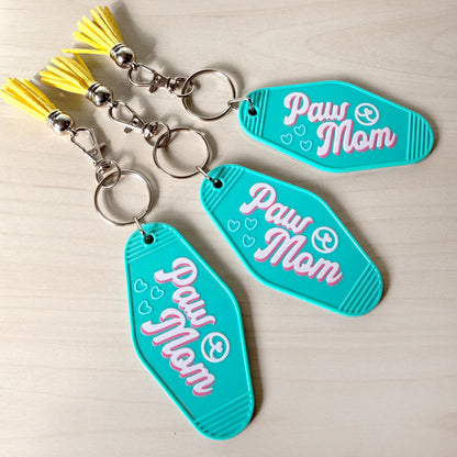 Turquoise 'Paw Mom' keychains with yellow tassels on a beige background