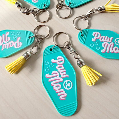 Turquoise keychains with 'Paw Mom' text and yellow tassels on a light wooden surface.