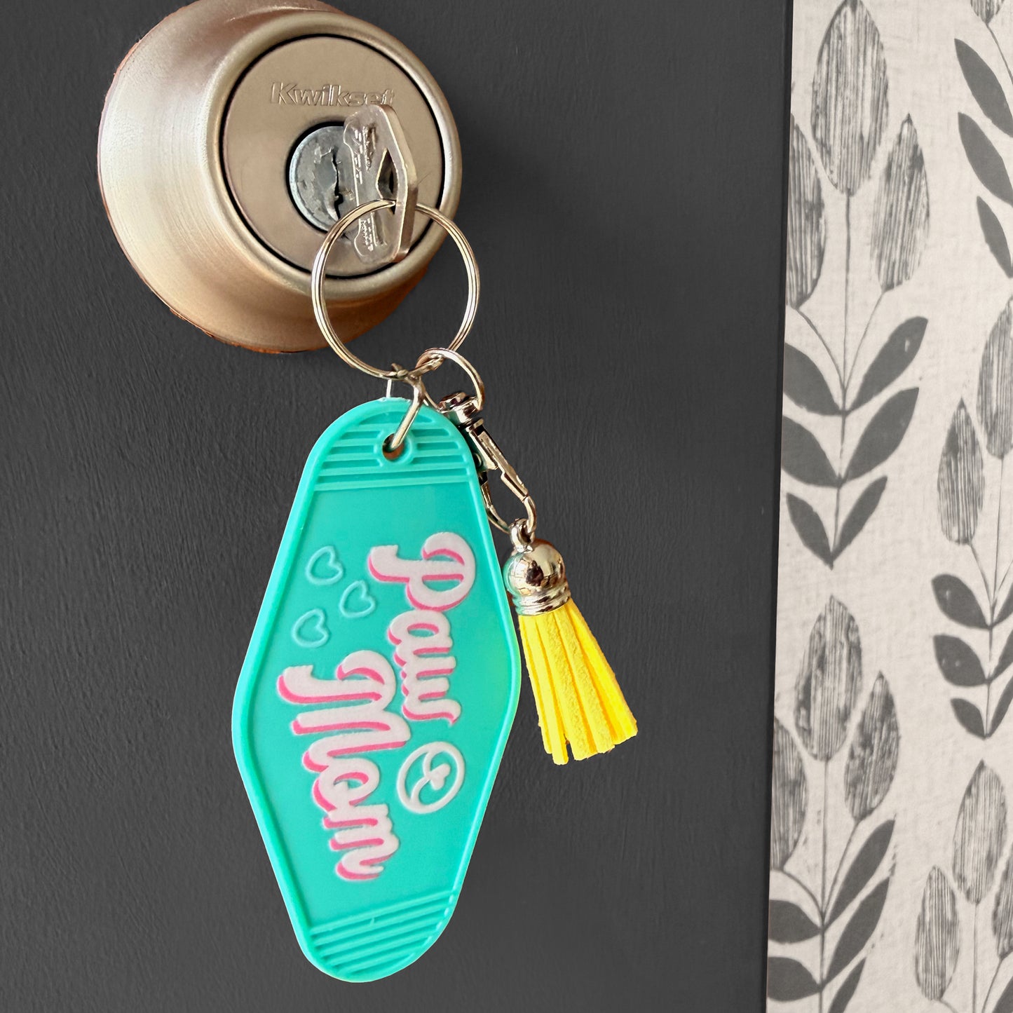 Turquoise keychain with pink text and yellow tassel attached to a door handle.
