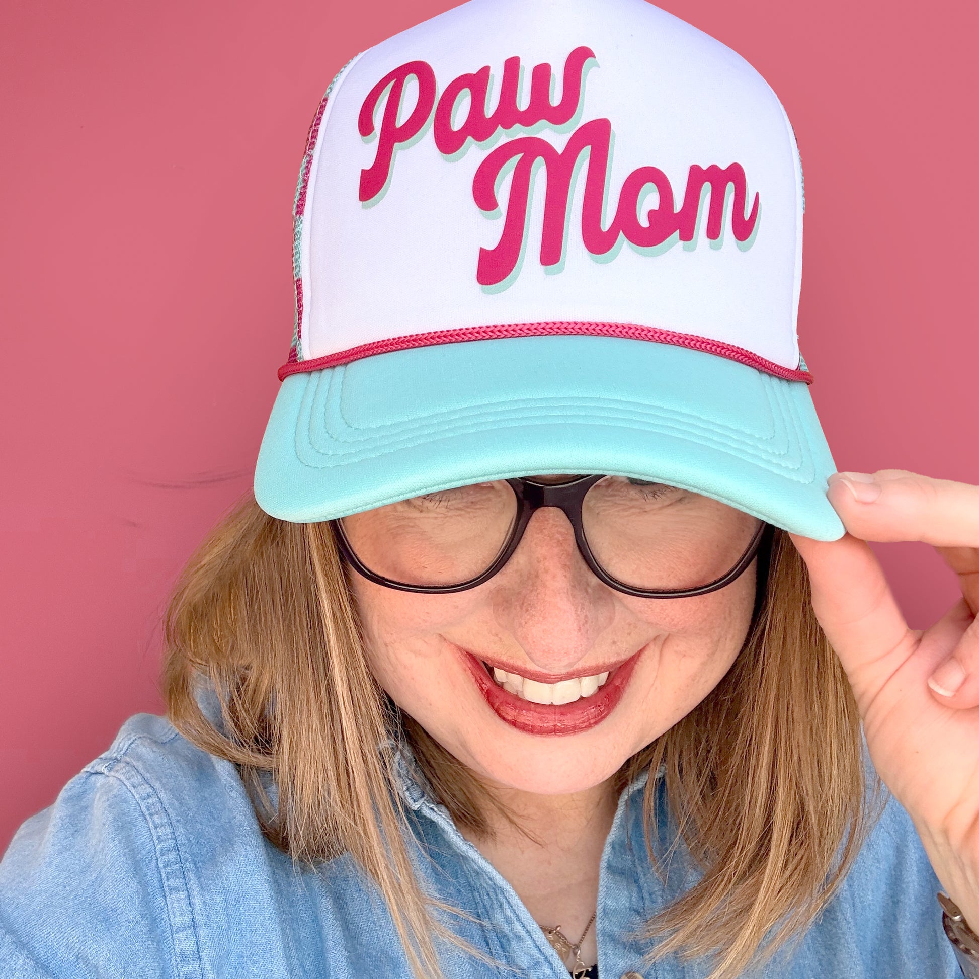 Person wearing a 'Paw Mom' cap with a pink and teal design against a pink background