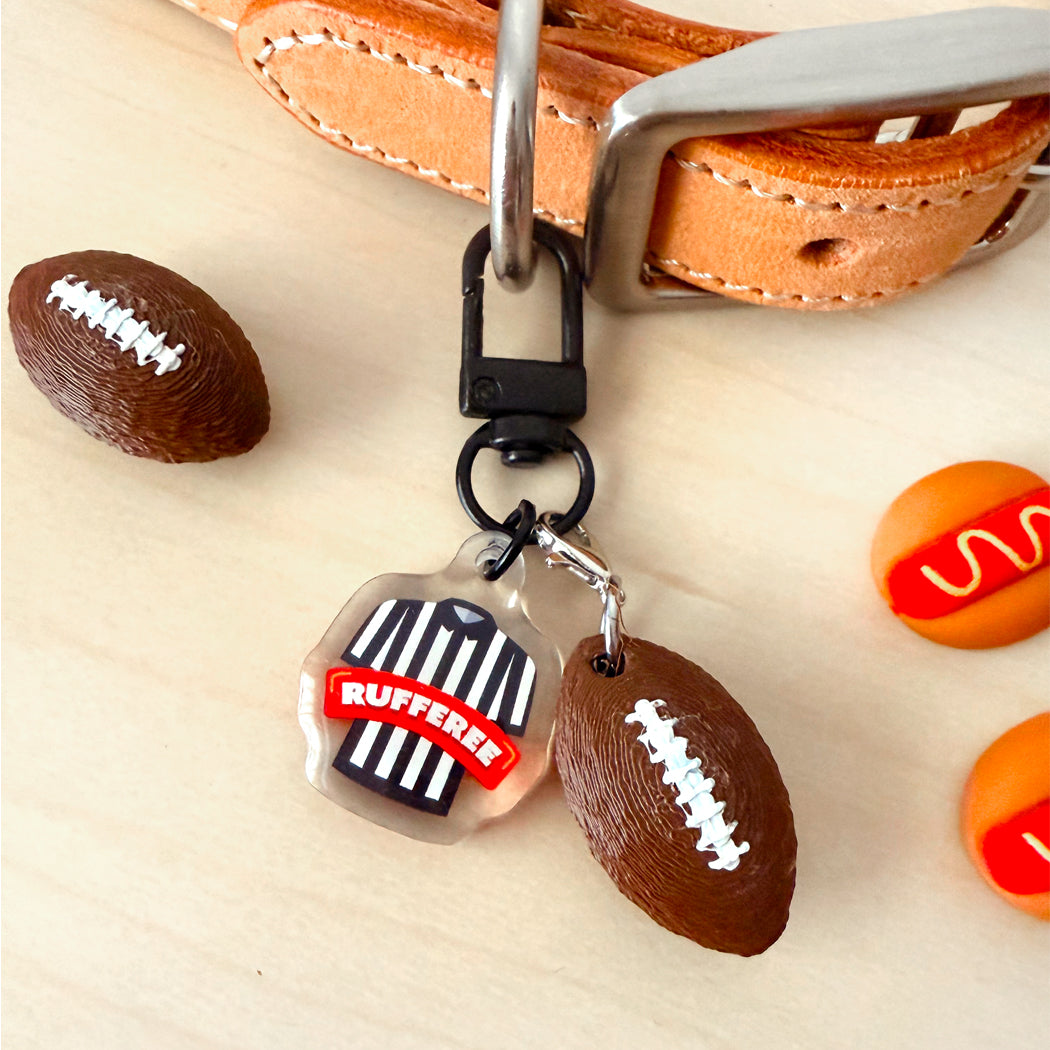 A close-up of a keychain or bag charm set on a light wood surface, featuring a clear shield-shaped charm with a referee shirt and the word "RUFFEE" in red, attached to a black clasp. The set also includes two football-shaped charms and a tan leather strap with a silver buckle in the background.