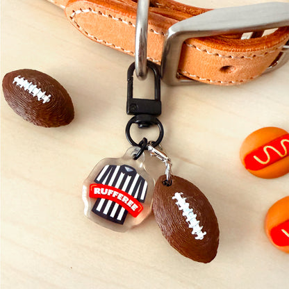 A close-up of a keychain or bag charm set on a light wood surface, featuring a clear shield-shaped charm with a referee shirt and the word "RUFFEE" in red, attached to a black clasp. The set also includes two football-shaped charms and a tan leather strap with a silver buckle in the background.