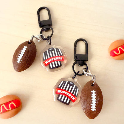 Two football-themed keychains on a light wood background. Each keychain has a black carabiner clip, a brown football charm with white laces, and a clear referee shirt charm with black stripes and a red banner reading 'RUFFEESE'.