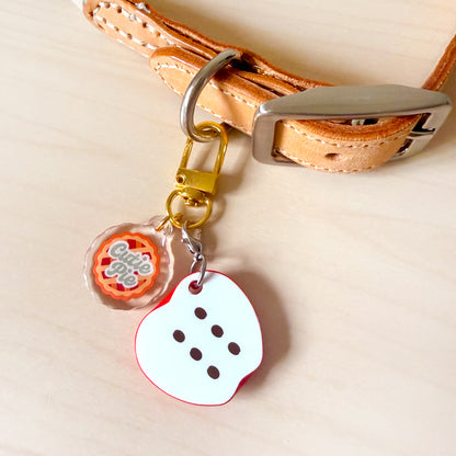 A close-up of a tan leather collar with a silver buckle, attached to a gold-tone key ring and split ring. Hanging from the ring are two charms: a clear acrylic round charm with an orange and white 'Chibi Cute' design, and a white die-cut heart-shaped charm with red trim and black dot details.