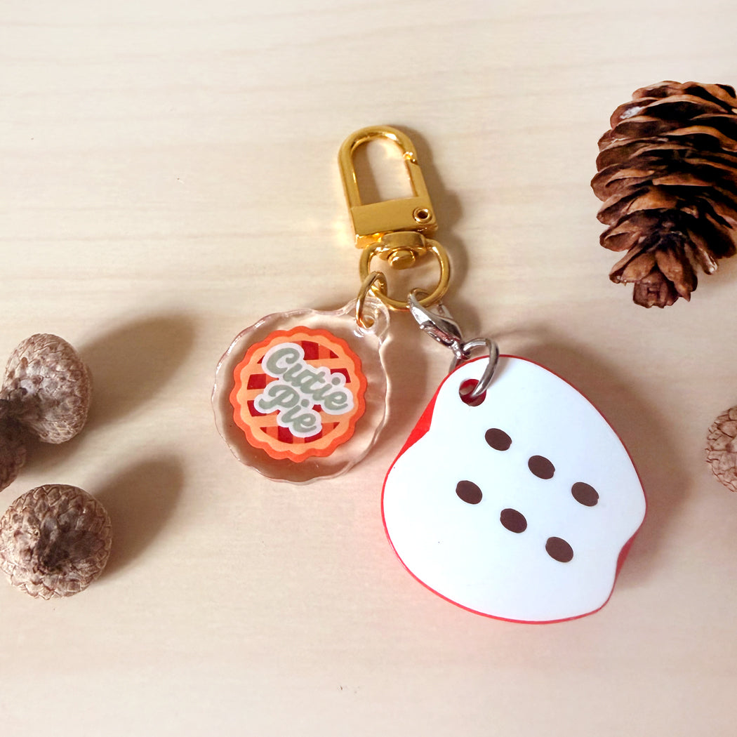 A keychain accessory set on a light wood surface, featuring a gold clasp with two charms: a round clear charm with an orange and gray 'Cutie Pie' design, and a white ghost-shaped charm with red trim and black dots.
