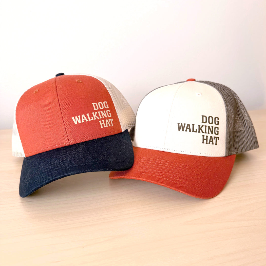 Two baseball caps with 'DOG WALKING HAT' text on a light wooden surface.