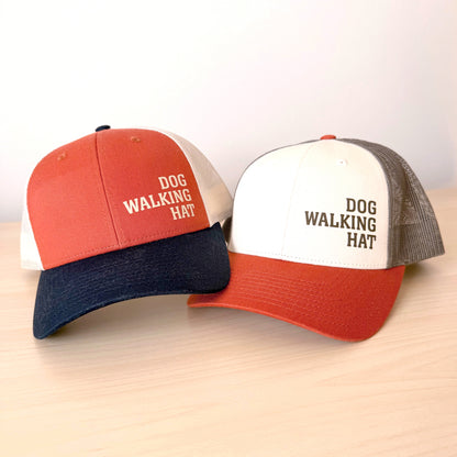 Two baseball caps with 'DOG WALKING HAT' text on a light wooden surface.
