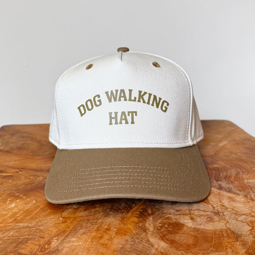 White and brown cap with 'DOG WALKING HAT' text on a wooden surface.