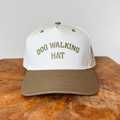 White and brown cap with 'DOG WALKING HAT' text on a wooden surface.