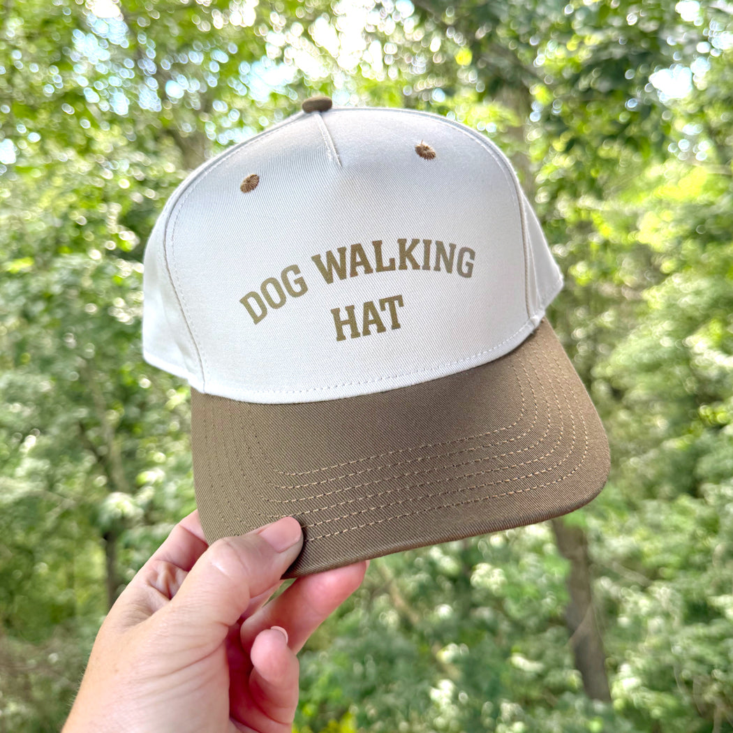 Cap with 'DOG WALKING HAT' text held in front of a blurred green outdoor background
