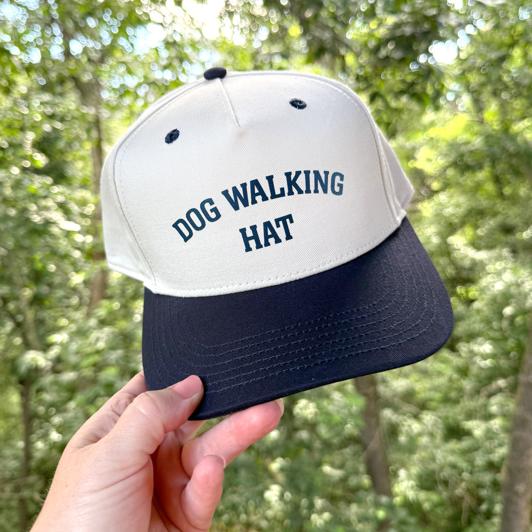 White and navy cap with 'DOG WALKING HAT' text held in front of a forest background