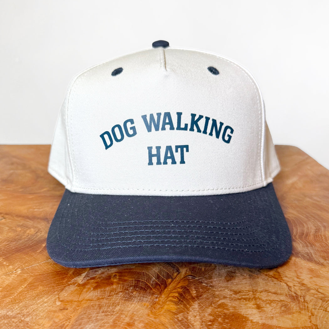 White and navy cap with 'DOG WALKING HAT' text on a wooden surface