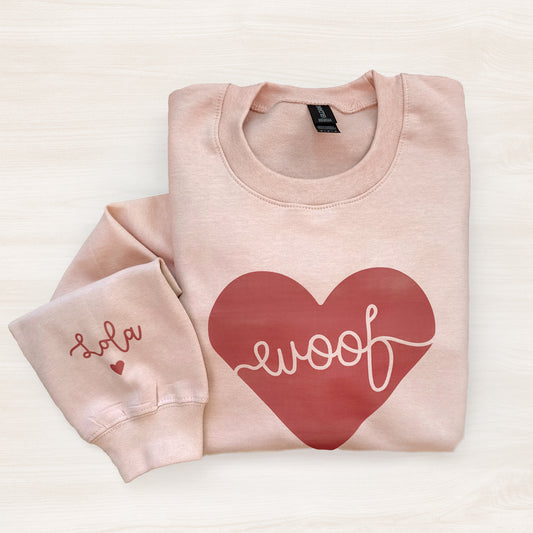 Folded pink sweatshirt with a red heart and 'woof' text on a light background